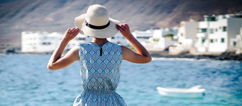 A girl in a beautiful dress holds a stylish hat in her hands and enjoys her leisure time, relaxing at sea. A lady stands with her back on the shore and looks at the ocean and the city on the horizon.