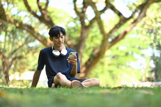 Relaxed young asian man with headphones using smartphone on grass in the park.