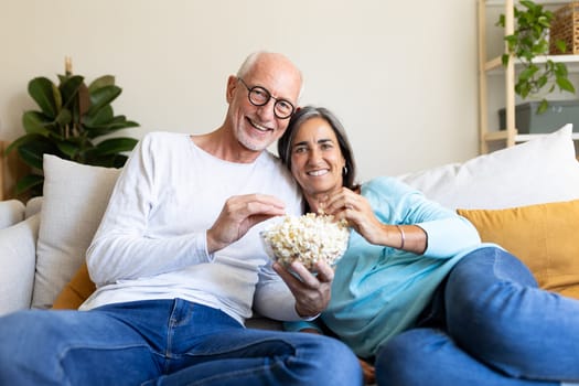 Mature happy married couple relaxing on the couch, watching a movie and eating popcorn. Looking at camera. Lifestyle concept.