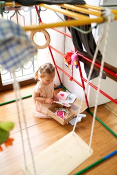 Little girl sits on the floor in a room near a swing and looks at postcards. High quality photo