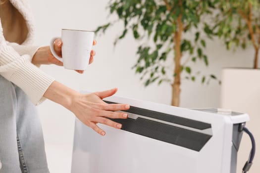 Woman warming hands near electric heater at home. Using heater at home in winter. Woman warming her hands. Heating season.