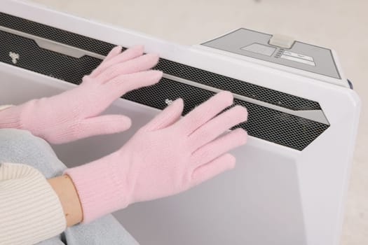Frozen young woman in mittens warming near radiator at home, closeup.