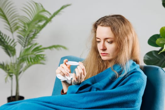 A young woman wrapped in a blue blanket sits on a couch, looking at a thermometer with a concerned expression. She appears to be unwell, surrounded by indoor plants.