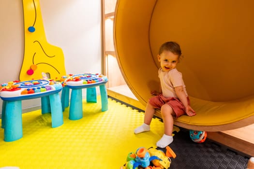 Smiling toddler sitting in a yellow play structure surrounded by colorful toys and activity tables in a vibrant playroom for fun and learning