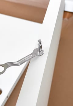 Assembling white furniture with metal wrench and cam lock fastener on brown surface.