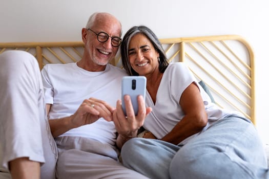 Happy mature caucasian couple looking at mobile phone together relaxing in bed. Lifestyle concept.