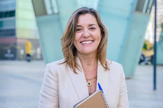 Confident middle-aged businesswoman smiling while holding a notebook. She stands outdoors in an urban setting with modern architecture in the background.