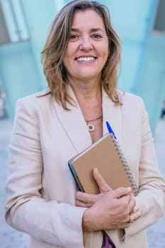 Confident middle-aged businesswoman smiling while holding a notebook. She stands outdoors in an urban setting with modern architecture in the background.