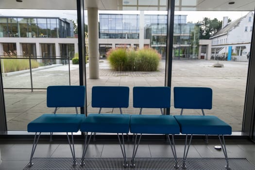 A row of four blue chairs are sitting in front of a window. The chairs are empty and the window is open, letting in a cool breeze. The scene is calm and peaceful, with the chairs