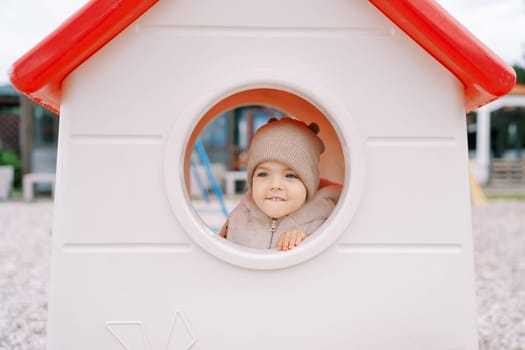 Little girl looks out the window of a toy house on the playground. High quality photo