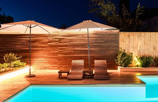 Elegant poolside area with lounge chairs and umbrellas at night. Modern outdoor design illuminated with warm lighting
