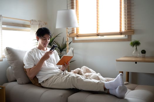 Carefree young adult man relaxing with a book and smartphone in cozy living room.