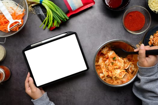 Overhead view of person holding digital tablet and mixing kimchi in stainless steel bowl on a dark countertop.