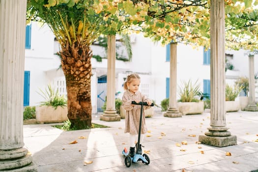 Little girl on a scooter rides between the columns of a pergola in the courtyard of a house. High quality photo