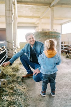Smiling dad with hay in hand squats next to little girl and looks at her. High quality photo
