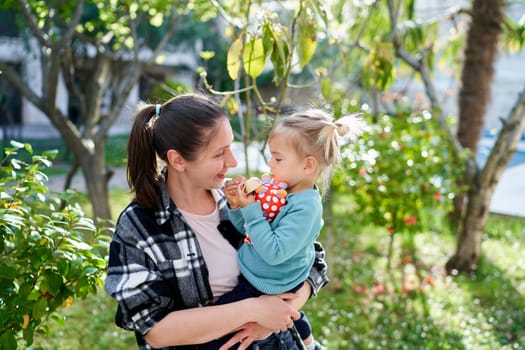 Smiling mother looking at little daughter sitting in her arms while standing in the garden. High quality photo