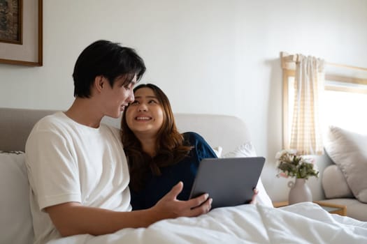 Loving adult couple using digital tablet under warm blanket in a cozy bedroom.
