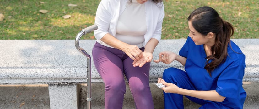 Caregiver or nurse giving and holding medicine pill to senior woman on park chair.