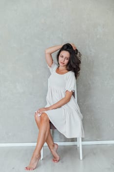 brunette in white dress poses sitting in a room on chair