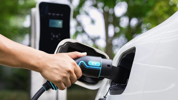 Hand insert EV charger and recharge electric car from charging station on nature and travel concept background. Technological advancement of alternative energy sustainability and EV car. Peruse