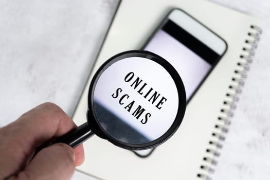A man's hand holds a black magnifying glass by the handle on a smartphone background with online scams text.