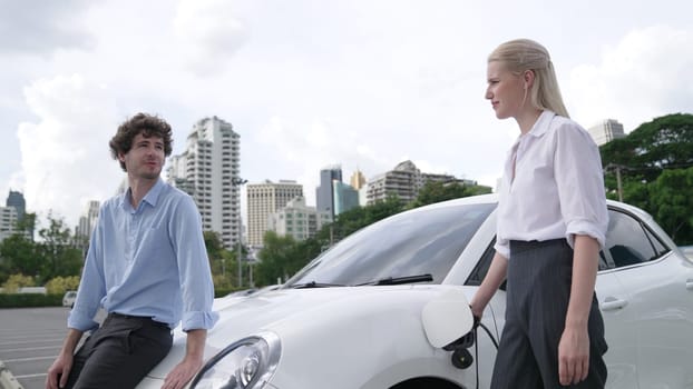 Progressive businessman and businesswoman install charger plug from charging station to electric car before driving around city center. Eco friendly rechargeable car powered by clean energy.