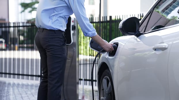 Progressive businessman install charger plug from charging station to his electric car before driving around city center. Eco friendly rechargeable car powered by sustainable and clean energy.