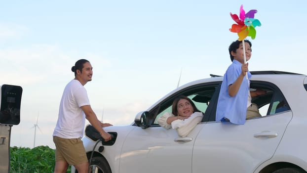 Concept of progressive happy family enjoying their time at wind farm with electric vehicle. Electric vehicle driven by clean renewable energy from wind turbine generator for charging station.