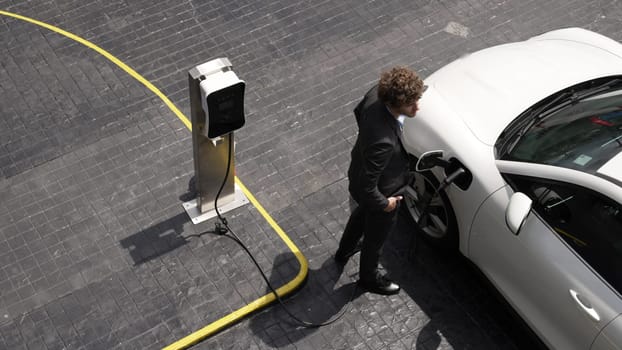 Progressive businessman install charger plug from charging station to his electric car before driving around city center. Eco friendly rechargeable car powered by sustainable and clean energy.