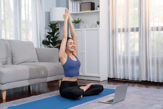 Asian woman in sportswear doing yoga exercise on fitness mat as her home workout training routine. Healthy body care lifestyle woman watching online yoga video on laptop. Vigorous