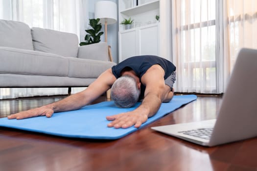 Senior man in sportswear being doing yoga in meditation posture on exercising mat at home. Healthy senior pensioner lifestyle with peaceful mind and serenity. Clout