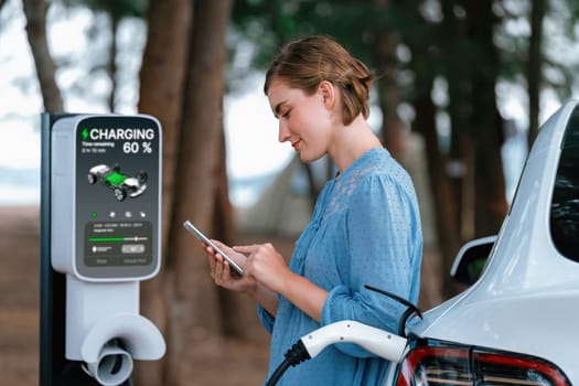 Holiday road trip vacation traveling to the beach camp with electric car, young woman checking battery from smartphone while recharge EV vehicle. Beach travel camping with eco-friendly car .Perpetual