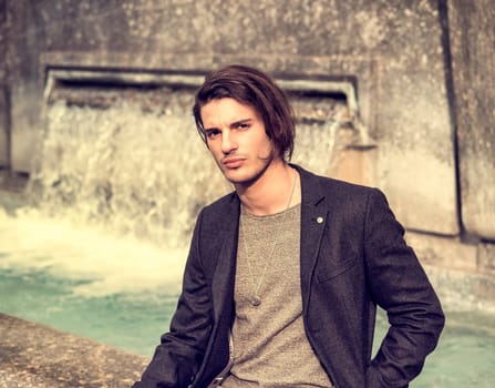 A man sitting on a rock next to a fountain. A young man enjoying the serenity of a city fountain, looking at camera