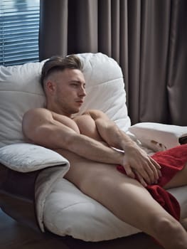 A man laying in a chair completely nude, hiding genitals with his hands
