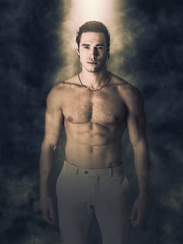 A man with no shirt standing in front of a dark background. Photo of a shirtless man posing against a dark backdrop with a dramatic light from above