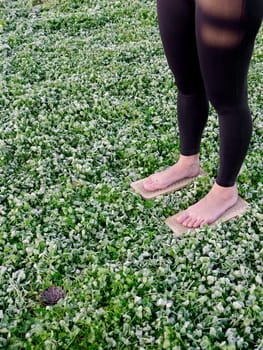 A barefoot woman stands on nails sadha board among the morning grass. Spiritual practice meditation in nature. Close-up, vertical