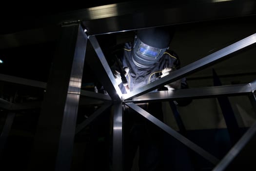Unrecognizable person in protective mask welding metal construction in dark room