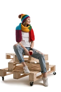Young pretty teenager in knitted yellow hat and scarf, white t-shirt and torn jeans sitting on wooden pallets with yellow knitted bag isolated shot