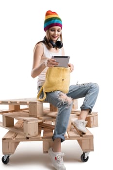 Young happy teenager in knitted yellow hat, white t-shirt and torn jeans sitting on wooden pallets with yellow knitted bag isolated shot
