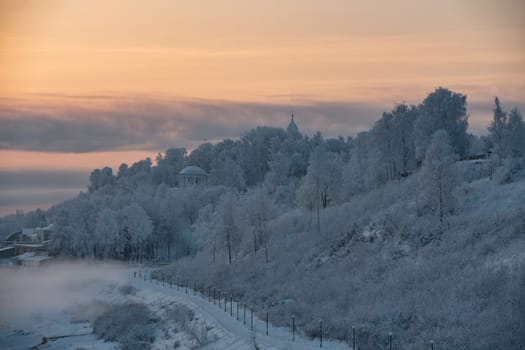 Beautiful Russian winter landscape with frozen river, embankment, street lamps and white rotund on a hill against pink and yellow sunset view