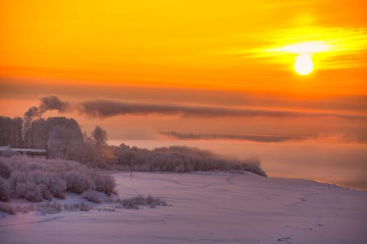 Beautiful Russian winter landscape with frozen river, bushes and houses against pink and yellow sunrise.