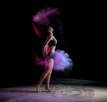 Side view of young woman shaking hair and throwing chalk while dancing in dark room with colorful illumination