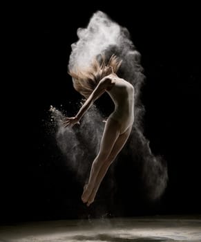 Slim girl wearing beige body jumping in white dust in dark room
