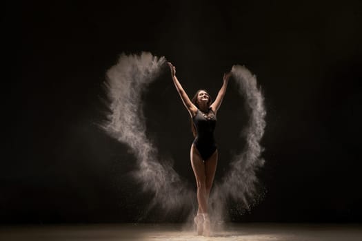 Slim ballerina in black bodysuit dancing with expression in flying white dust on white background