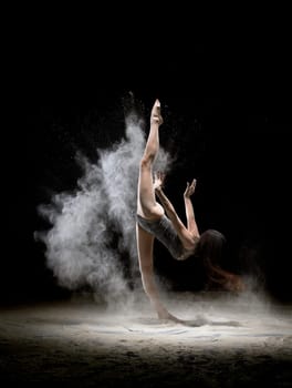 Side view of sportive dance in black bodywear doing splits while standing and dancing in white flying dust on black background