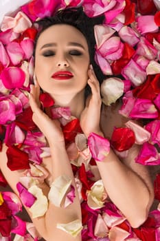 Attractive female with bright makeup keeping eyes closed and touching face while lying in water covered with delicate rose petals