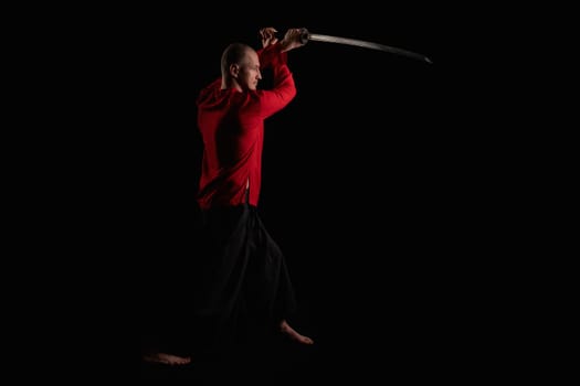 Man wearing red shirt doing exercises with a sword full-length profile shot in the dark room