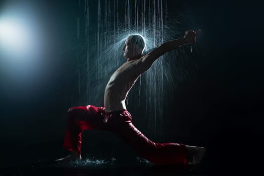 Shirtless man in red trousers doing yoga under the rain in the dark