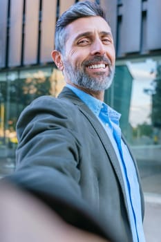 Mature businessman with gray hair and beard taking a selfie in a modern urban setting, standing near a glass office building in daylight.