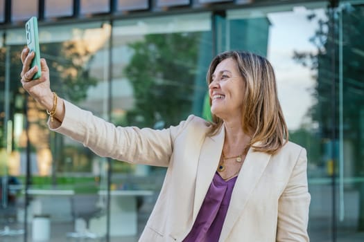 Mature businesswoman with gray hair and beard taking a selfie in a modern urban setting, standing near a glass office building in daylight.